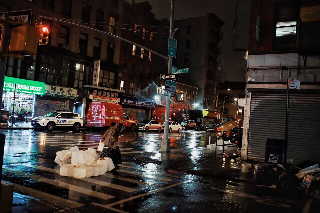 building exterior, architecture, city, night, built structure, street, illuminated, wet, transportation, group of people, water, city life, building, real people, women, rain, mode of transportation, city street, road, outdoors, rainy season