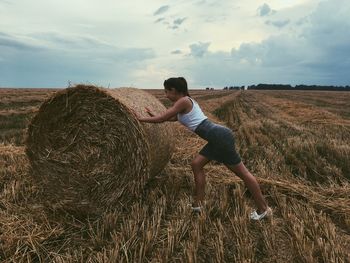 Side view of young woman pushing hay bale on grassy field against sky during sunset