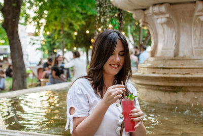 Beautiful young woman drinking strawberry mojito cocktail in park in city