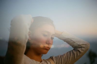 Close-up portrait of young woman with arms raised against sky