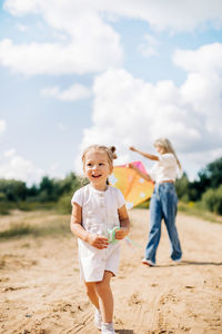 A little girl in a white dress is running along the road with a string of a kite in her hand