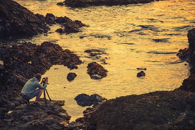 High angle view of man photographing on rock at beach during sunset