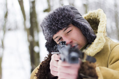 Soldier in brown old coat and fur hat aims with a weapon in the forest in winter