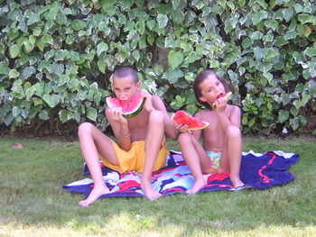 Full length portrait of shirtless siblings eating watermelons against plants on grassy field