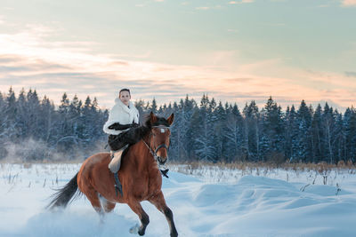 A girl in a white cloak rides a brown horse in winter.