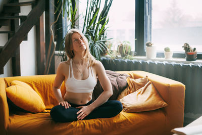 Serene calm girl sitting on the sofa in the lotus position, doing yoga exercises for relaxation