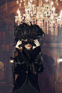 Portrait of young woman wearing mask while standing below illuminated chandelier