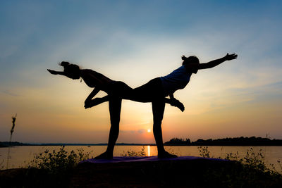 Silhouette friends doing yoga at beach against sky during sunset