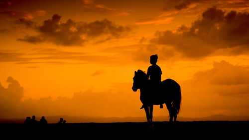 Silhouette boy riding horse against orange sky