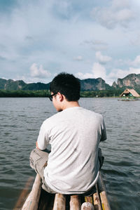 Rear view of man sitting on wooden raft over lake against sky