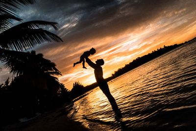 Silhouette man playing with child at beach against sky during sunset