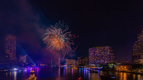 Firework display over river by buildings against sky at night