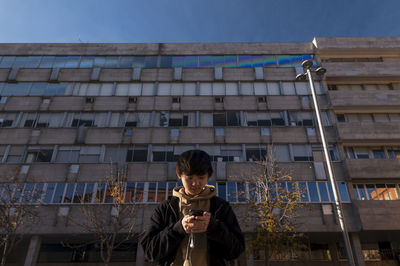 Asian teenage boy looking at mobile phone against building on street. madrid. spain