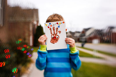 Boy covering face with handprint on paper against sky