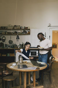 Female architect analyzing blueprint by male colleague having lunch in home office