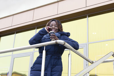 Low angle view of woman holding coffee cup talking on phone against building