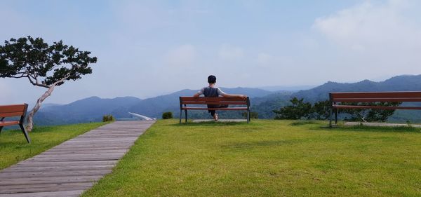 Rear view of man sitting on bench on field against sky