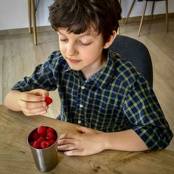 Cute boy eating raspberries while sitting on chair at home