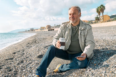 Man listening music or podcast in headphones and drinking coffee while sitting on winter  beach.