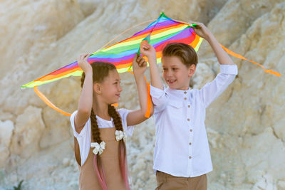 A girl and a boy are carrying a bright kite with their hands raised high above their heads.