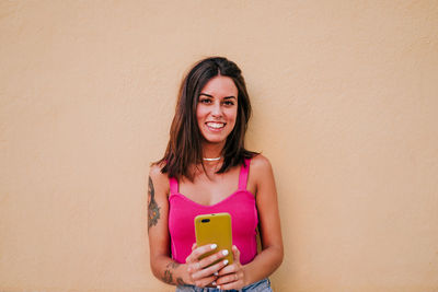 Portrait of smiling young woman holding mobile phone against wall