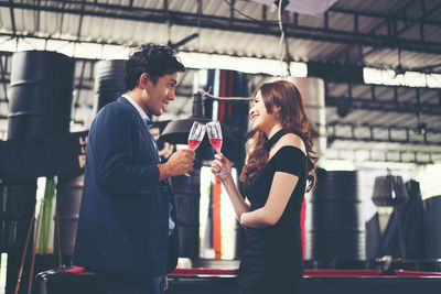 Side view of smiling couple toasting red wineglasses while standing in bar