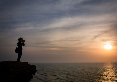 Silhouette woman photographing sunset while standing on rock formation by sea against cloudy sky