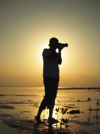 Silhouette of man standing on sea shore against sunset sky