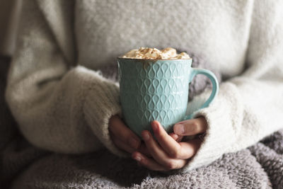 Woman covered with blanket holding mug of hot drink with whipped cream