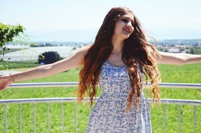 Smiling woman with arms outstretched standing by railing against field on sunny day