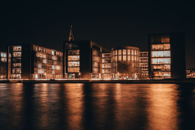 Illuminated buildings by river against sky at night