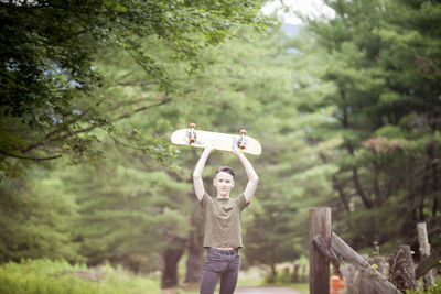 Portrait of teenage boy holding skateboard while standing against trees