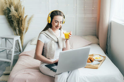 Cheerful woman holding juice while using laptop at home