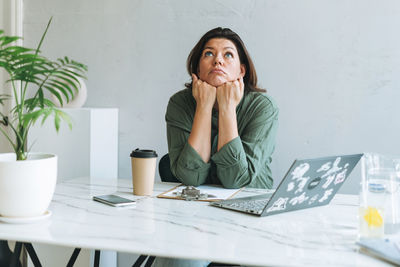 Young thinking unhappy brunette woman plus size working at laptop on table with plant in office