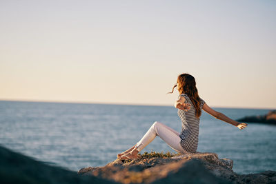 Full length of young woman sitting on rock against sea