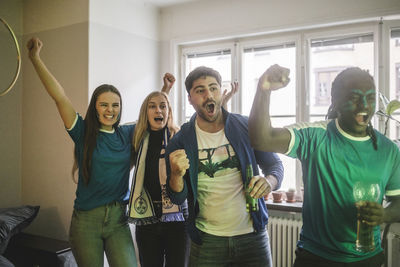 Young cheerful friends watching soccer match while standing in living room