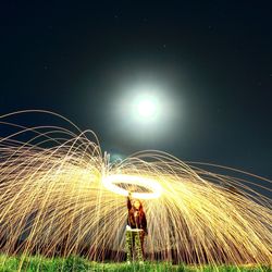 Woman spinning wire wools while standing on grassy field against sky at night