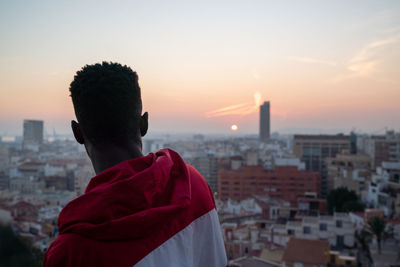 Side view of confident young ethnic male in trendy outfit and true wireless earphones admiring picturesque view of coastal city against cloudless sunset sky