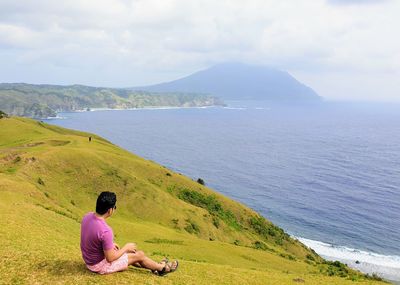 Full length of man looking at seascape while sitting on mountain