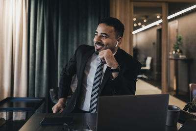 Smiling businessman looking away while talking through wireless in-ear headphones in board room