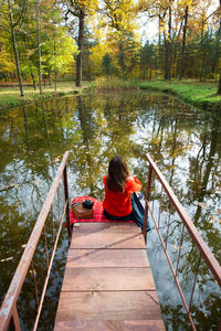 Rear view of woman sitting on footbridge over lake in forest