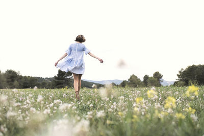 Full body back view of female in short blue dress walking through meadow covered with grass and flowers next to forest and mountain valley under cloudless sky in daytime