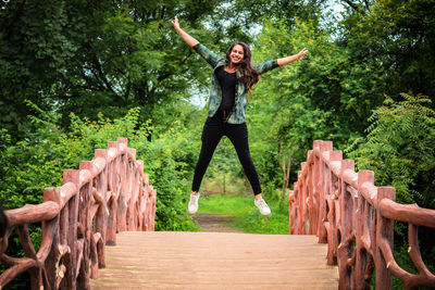 Portrait of smiling young woman with arms raised jumping on footbridge