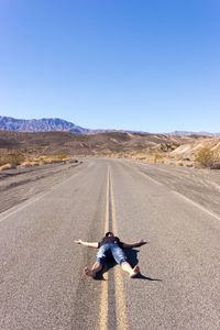 Full length of woman lying on country road against clear blue sky during sunny day