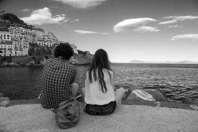 Rear view of man and woman sitting by sea against sky in town
