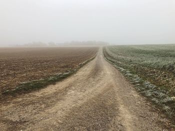 Dirt road amidst field against sky during foggy weather