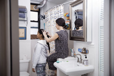 Mother sink tying daughter's hair while sitting on sink in bathroom