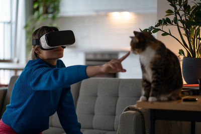 Positive casual woman owner in modern vr headset pulls hand to cat sits on sofa of apartment