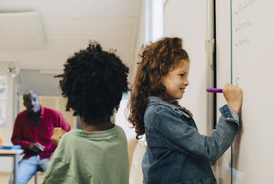 Boy standing by girl writing on whiteboard in classroom