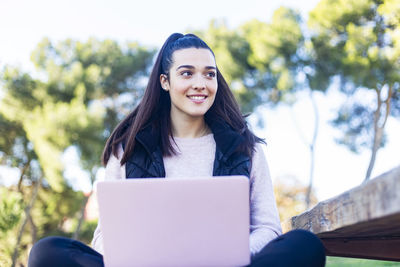 Low angle view of smiling young woman with laptop looking away while sitting in park